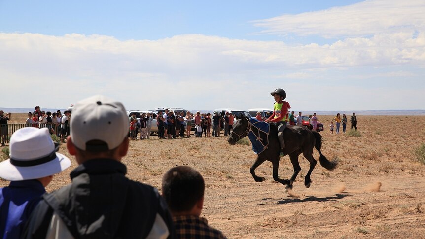 A child jockey races in Mongolia as crowds stand and watch from the sidelines.