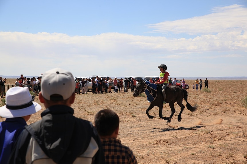 A child jockey races in Mongolia as crowds stand and watch from the sidelines.