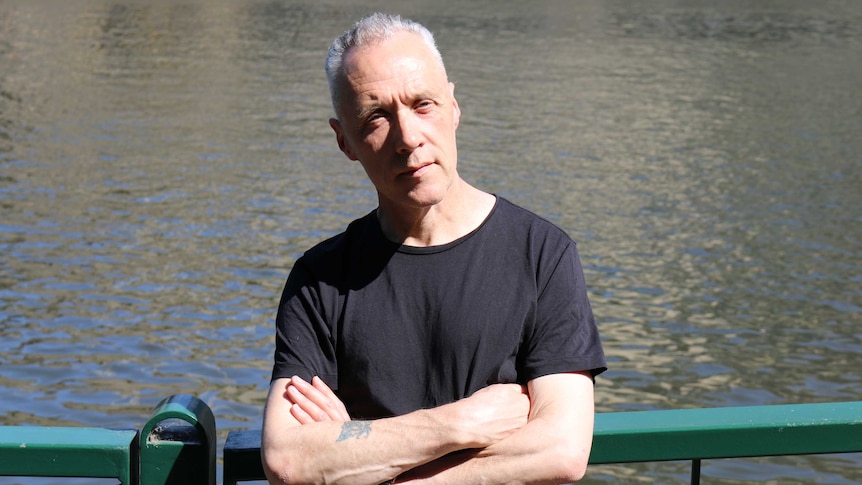 Before a backdrop of water, a man stands against a green railing in black t-shirt, with arms folded, looking forward.