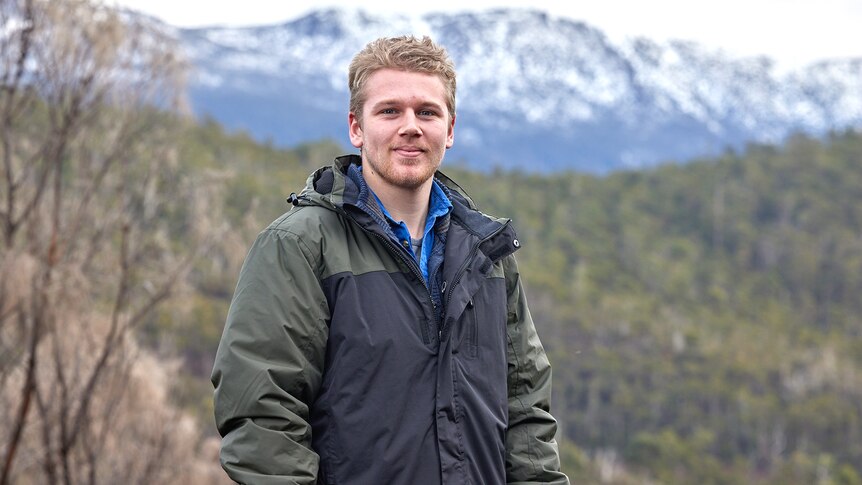 A man with blond hair wearing a coat in front of a forest and mountains