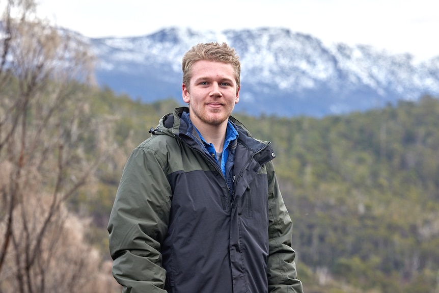 A man with blond hair wearing a coat in front of a forest and mountains