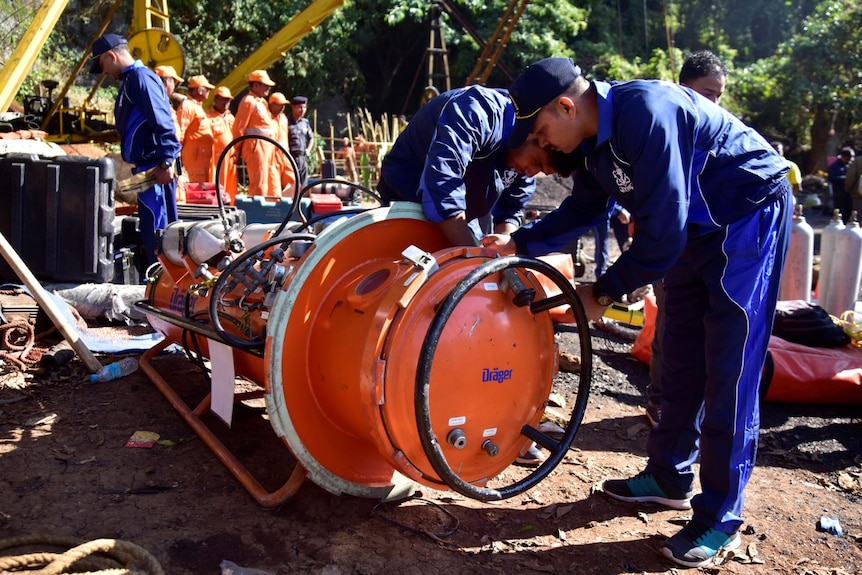 Men in blue uniforms inspect a large, orange piece of machinery.