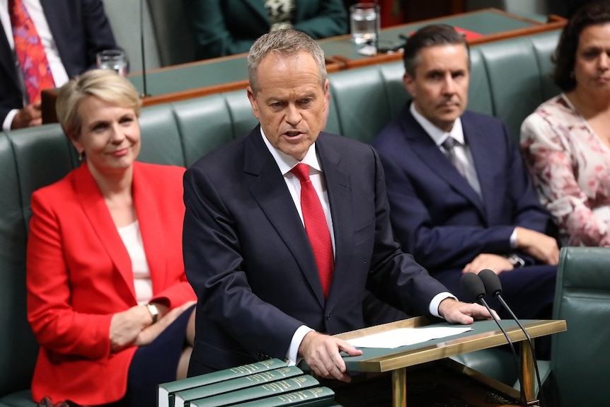 A man in a suit stands as he speaks from a lectern, while others sit along benches behind him.