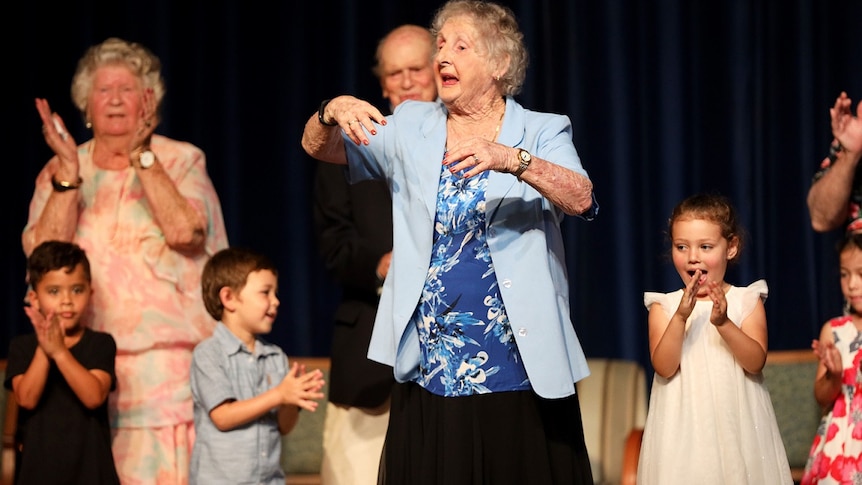 An elderly woman in a blue blazer dances next to children.