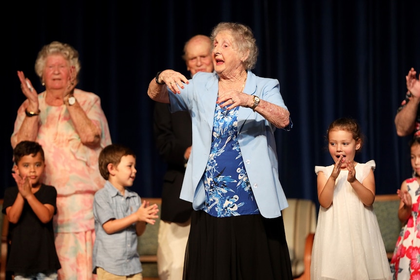 An elderly woman in a blue blazer dances next to children.