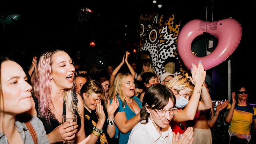 A group of young women and non-binary people are smiling and having fun while gathered on a dance floor and looking up. 