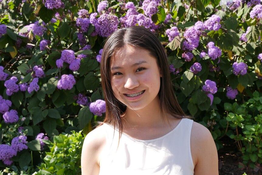 A teenage girl stands in front of a bush with purple flowers and smiles. She wears a white singlet and has braces.