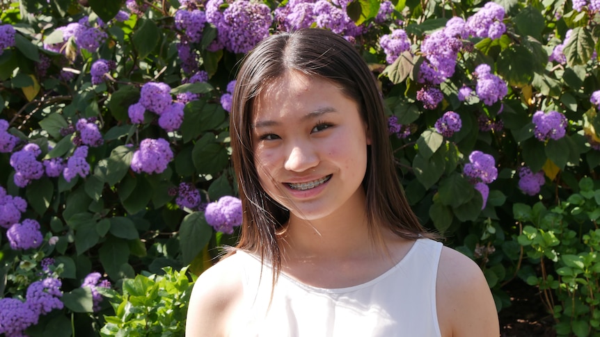 A teenage girl stands in front of a bush with purple flowers and smiles. She wears a white singlet and has braces.