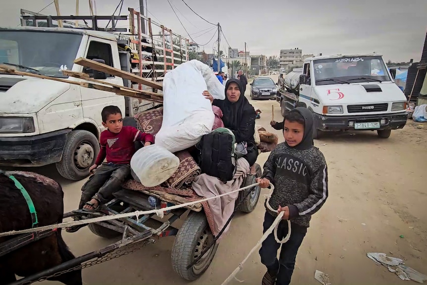 A woman sits on a cart pulled by a donkey and stacked with belongings, while children walk alongside