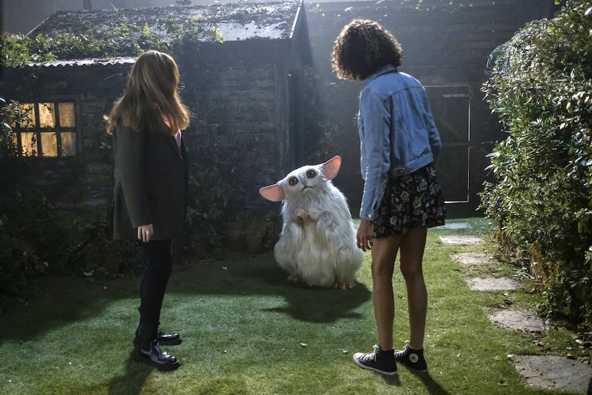 Two woman stand in a backyard with their backs turned, staring at a small white creature a bit like a furby