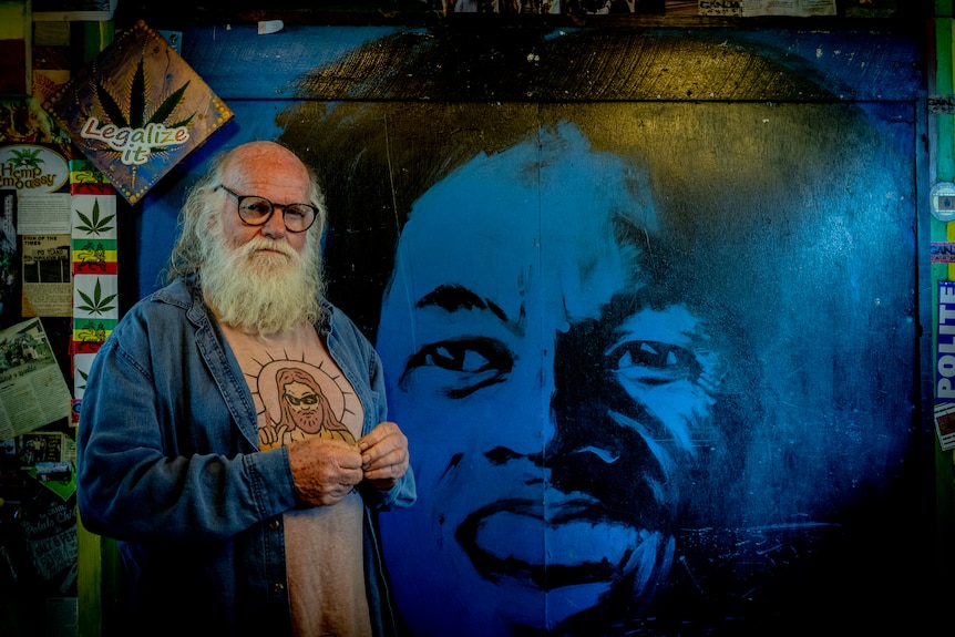 An older man with a white beard and hair, bald on top, wearing glasses stands next to a mural of a large blue face