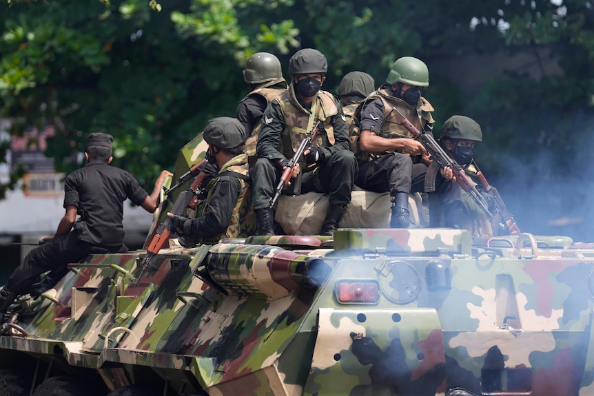 Six soldiers holding guns sit on top of an armoured vehicle dressed in camouflage.