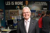 Les Murray stands in the office with a promo poster for Les is More on the back wall.