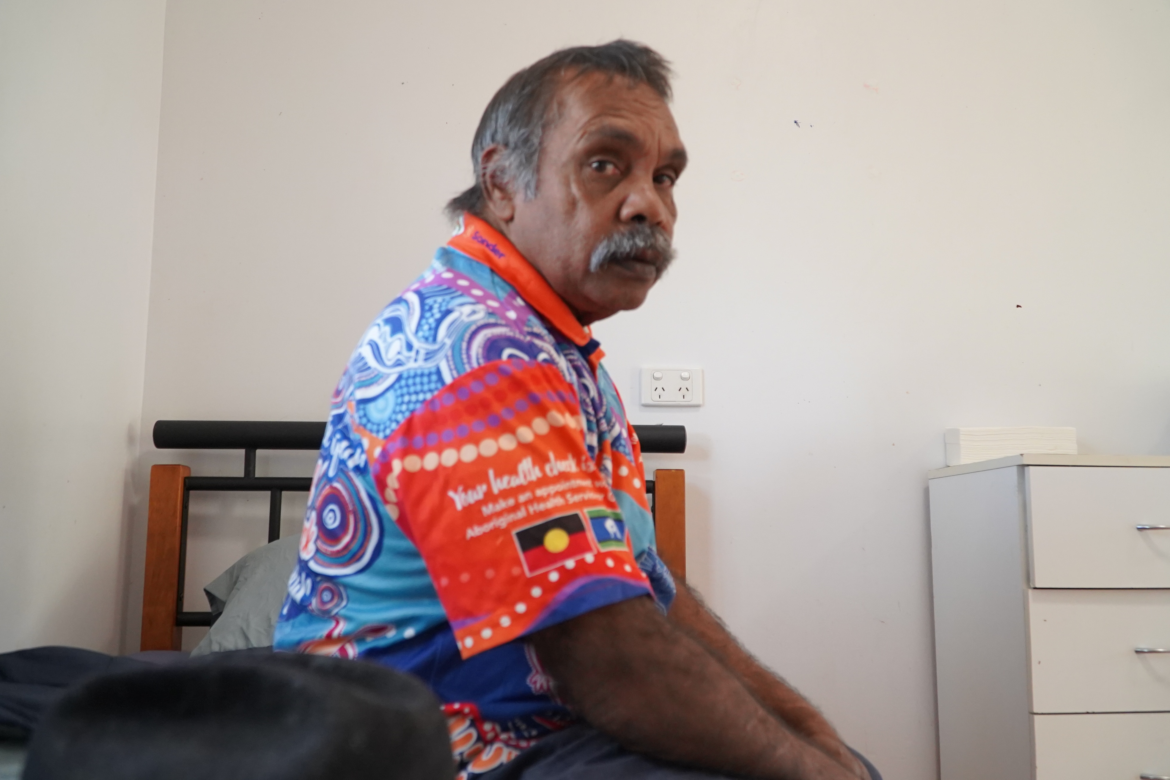 Tim McNamara, an Indigenous man, sits on a bed and looks right past his shoulder to the camera.