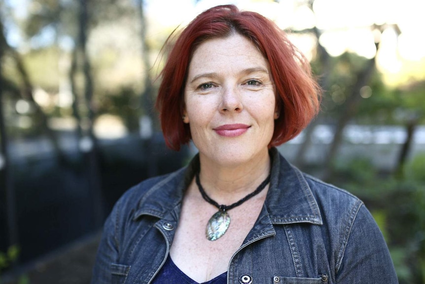 A woman with intensely coloured hair, wearing a dark denim jacket, stands outside.