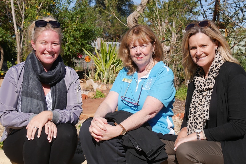 Three blonde women smile as they sit on a low wall in front of a native garden on a sunny day.