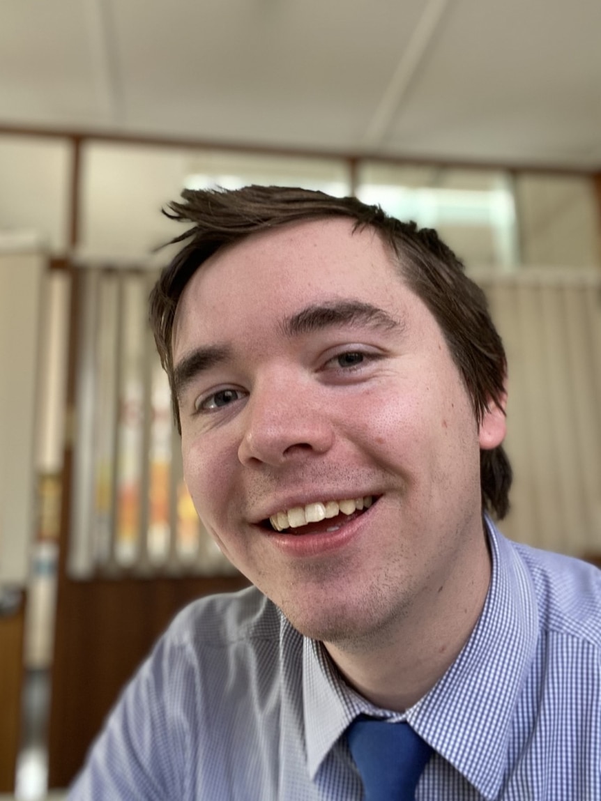 A young man with brown hair in a light blue checkered shirt smiles at the camera