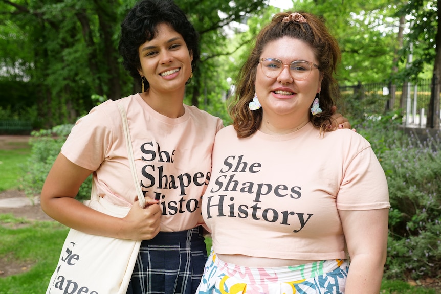 Two women wear shirts that say She Shapes History on them, both are smiling in a park. 