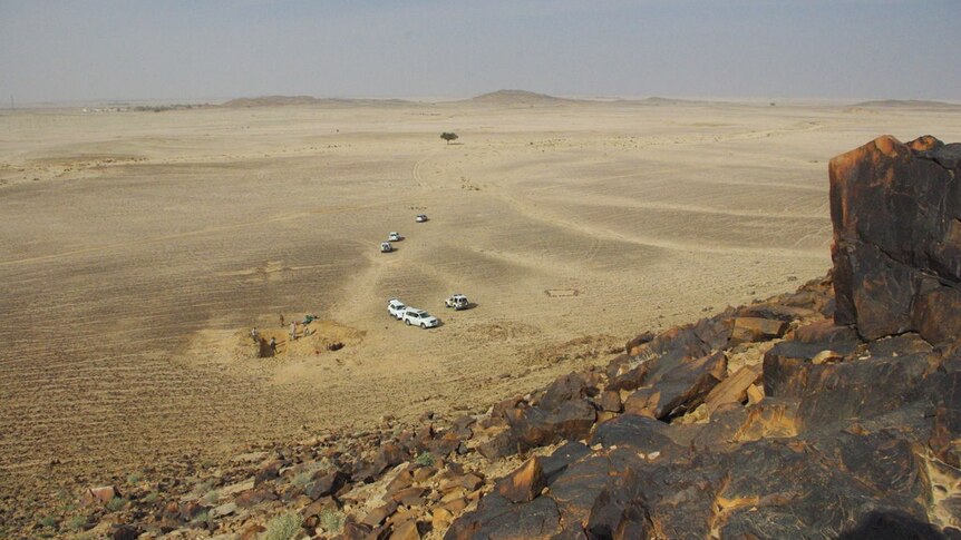 Cars and people digging can be seen in the desert from the top of a rocky hill.