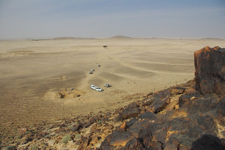 Cars and people digging can be seen in the desert from the top of a rocky hill.