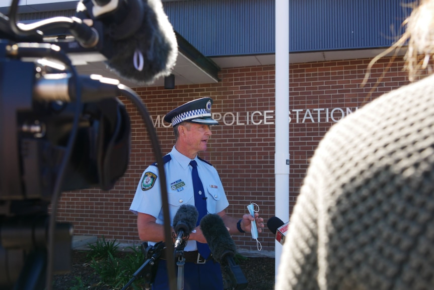 Man in police uniform speaks to microphones in front of police station.