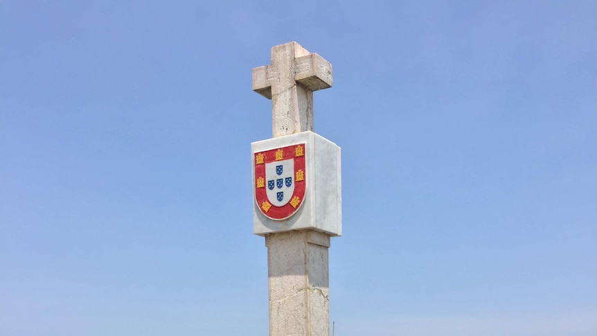A tall pillar with a cross at the top stands on a hill overlooking trees and the ocean.