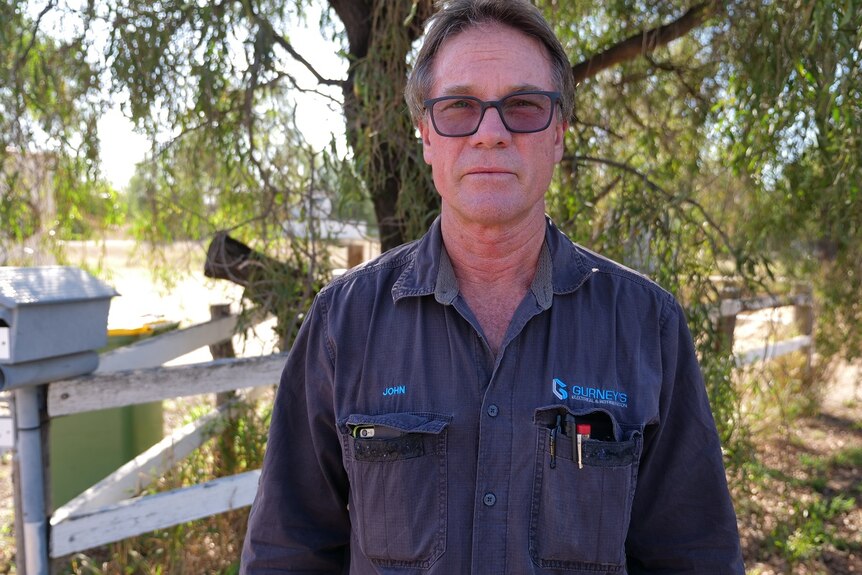 John Gurney, wearing glasses, stands in front of his mailbox and a tree, wearing a longe sleeve, dark blue button up shirt.