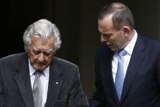 Bob Hawke holds a walking cane in a doorway as he stands next to Tony Abbott who offers a hand