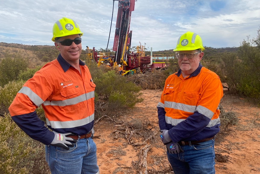 Two men stand in front of a drill rig smiling, outback scrub behind them