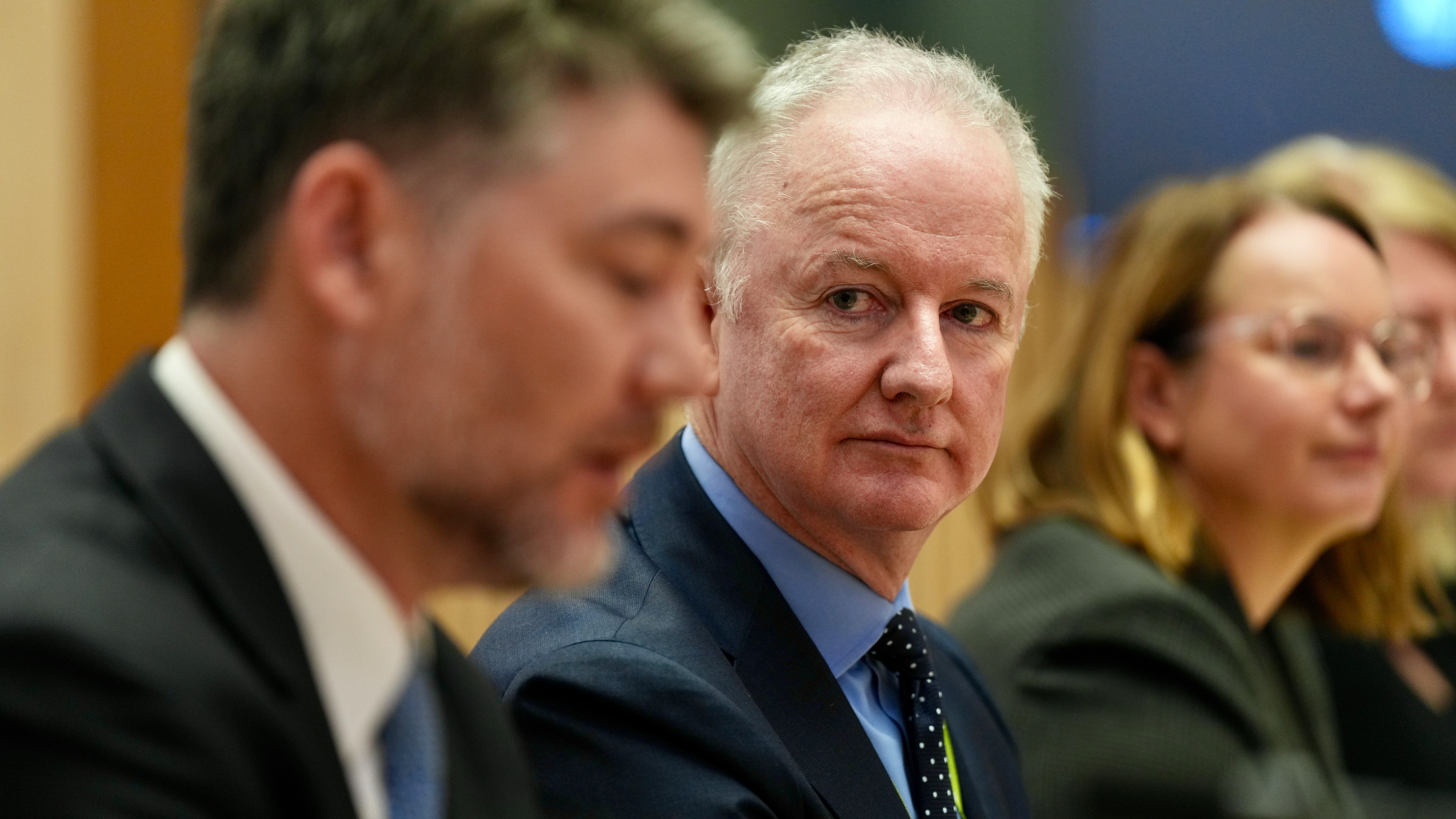 A grey-haired man (Hugh Marks) wearing a suit is in focus, with blurred smartly dressed people sitting either side.