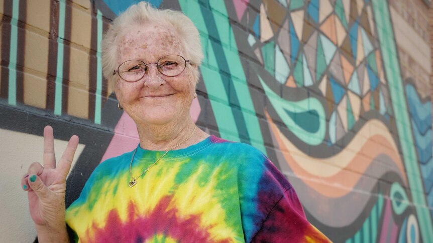 An older woman standing in a bright t-shirt with her hands in a peace sign. She is standing in front of a colourful wall.