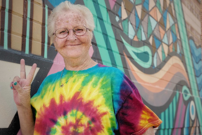 An older woman standing in a bright t-shirt with her hands in a peace sign. She is standing in front of a colourful wall.