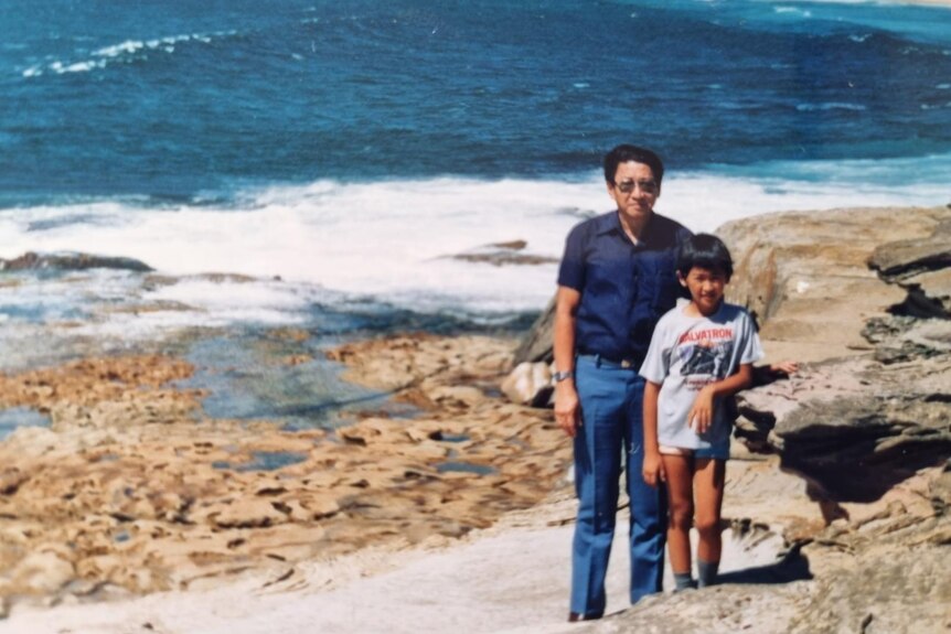A young boy and his father stand on rocks in front of the ocean.