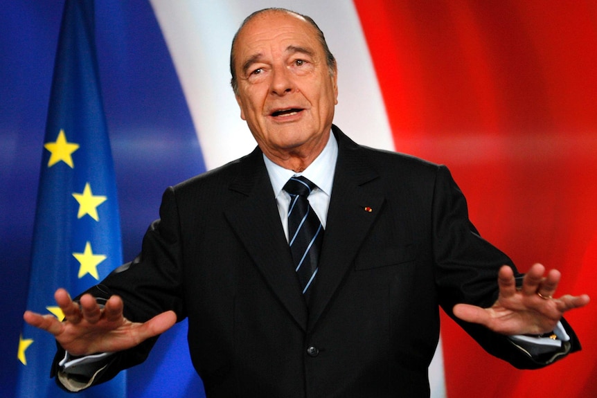 French president Jacques Chirac puts his hands out in front of him as he stands in front of a French flag.