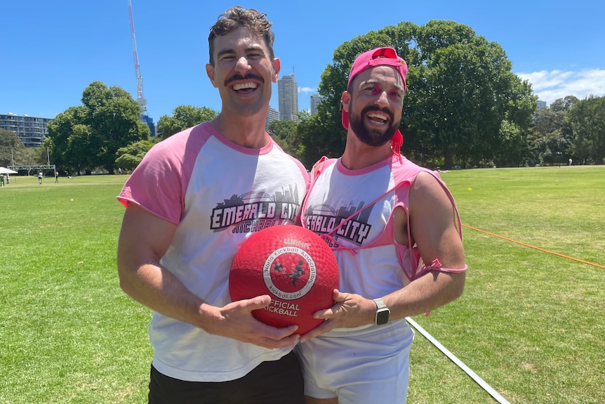 Two men in pink and white sports tops hold a big red ball and laugh 
