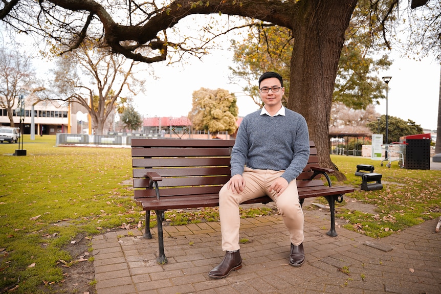A man in a knitted sweater and glasses sits on a park bench, smiling.