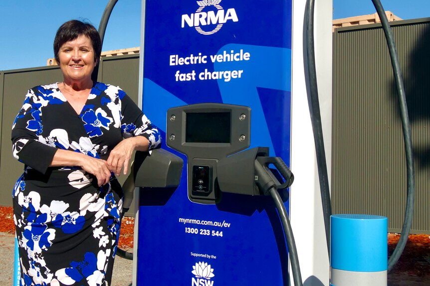 a white woman with black hair and a blue dress standing next to an electric vehicle charging station