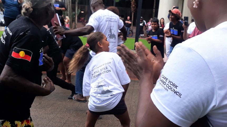 A young girl swings her hair, while others in matching T-shirts dance in the courtyard