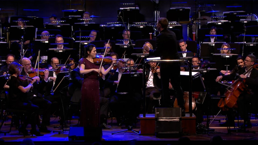 Emily Sun plays violin standing on stage in front of an orchestra. Her eyes are closed and she wears a long maroon beaded gown.