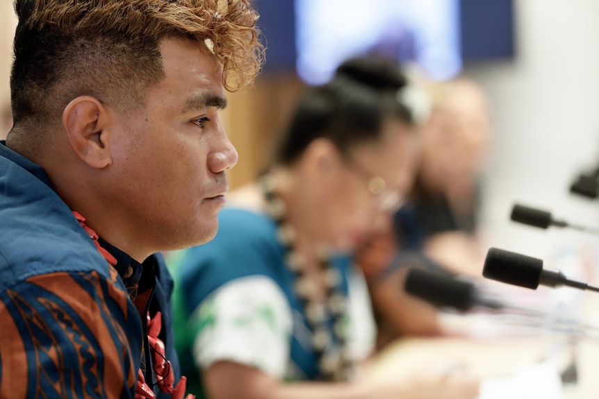 A side-on profile of a Samoan man sitting at a desk with microphones.