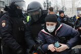 A woman wearing a beanie and face mask is gripped by several police officers in winter coats and helmets.