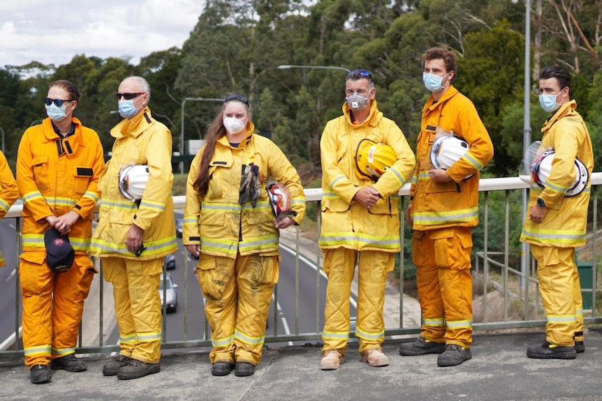 Volunteer firefighters in yellow suits stand sadly on the bridge.