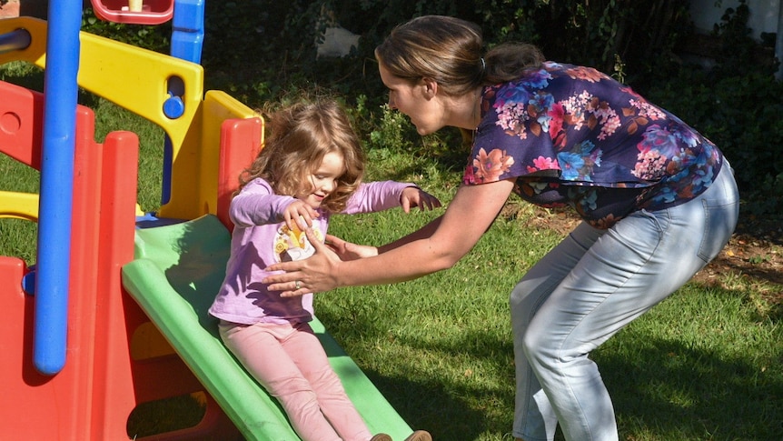 A small girl slides down a slippery dip as her mother wearing a floral top catches her.