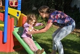 A small girl slides down a slippery dip as her mother wearing a floral top catches her.