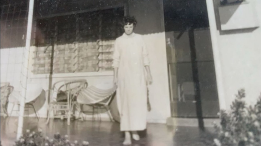 A grainiy black and white picture of a young tall woman in a light-codressing gown on the verandah of her old house in Wittenoom