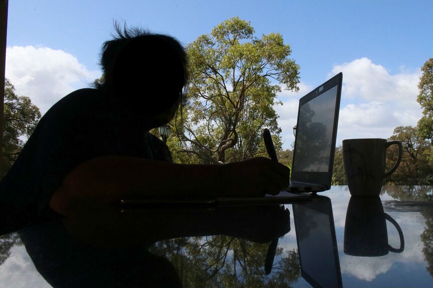 A woman silhouetted against the background sits down at a table outdoors with a laptop and a coffee cup.