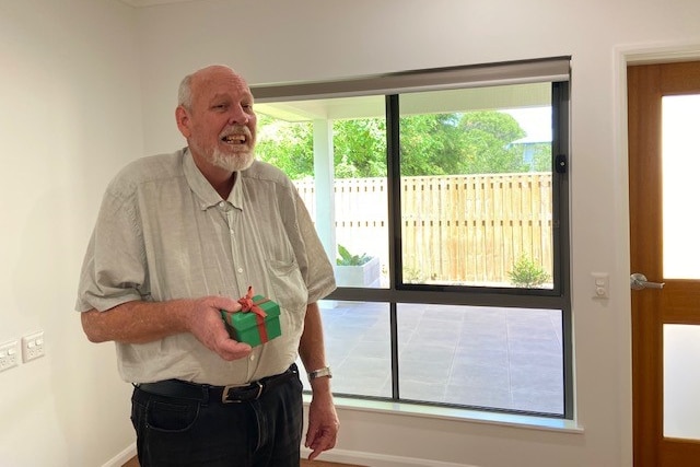 a man in a white shirt standing in a room smiling, holding a small present box