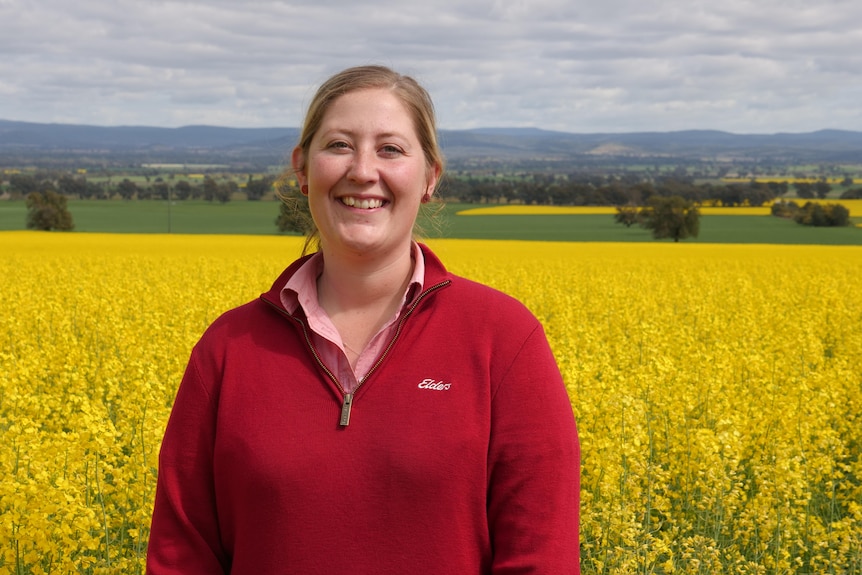 A young woman wearing a red jumper standing in front of a canola field 