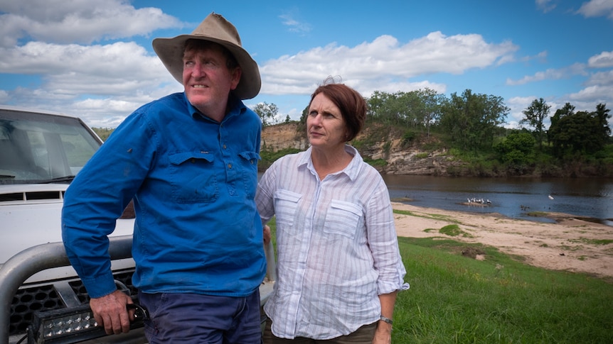 A man wearing an old hat and a woman lean against a ute parked on their property with a river scene in the background.