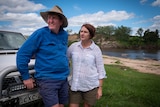 A man wearing an old hat and a woman lean against a ute parked on their property with a river scene in the background.